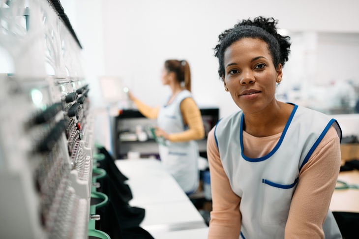 Sanft lächelnde Person in hellblauer Arbeitskleidung steht vor industrieller Stickmaschine, im Hintergrund verschwommen weitere Person