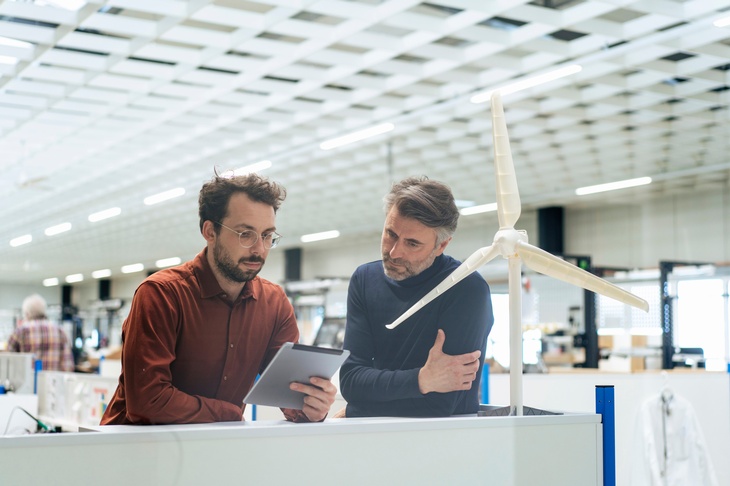 Personen blicken auf ein Tablet und lehnen sich an eine Front auf dem ein Modell einer Windturbine in einem Werksraum steht